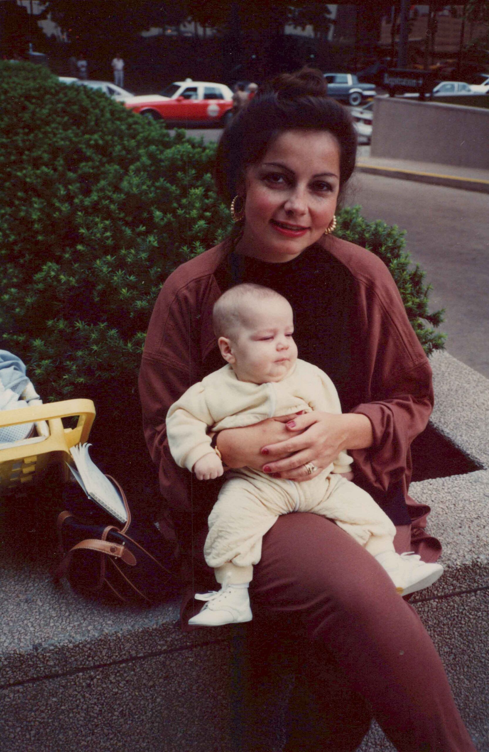 Woman in brown outfit sitting on a curb, holding a baby in cream clothing with a green hedge behind them and cars in the background.