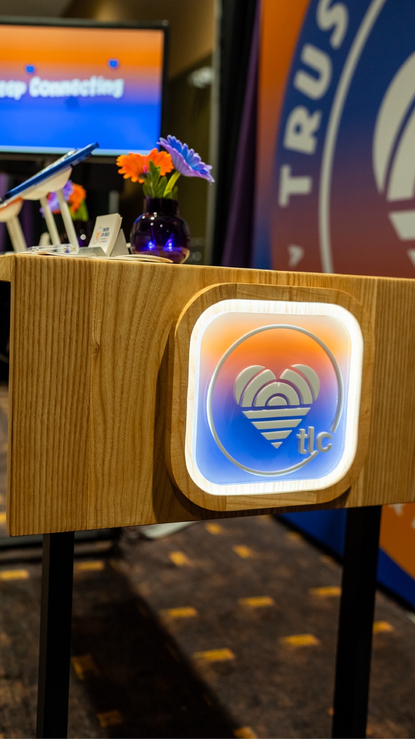 Wood podium with a glowing circular TLC logo in blue, orange, and pink, and a small vase of orange and purple flowers on top while a screen is blurred in the background.