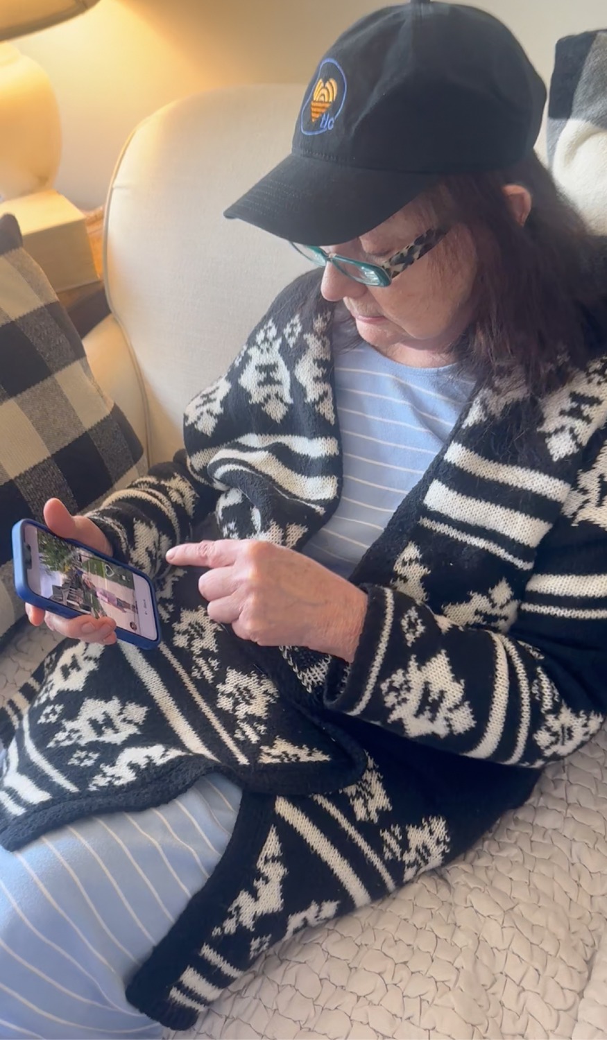 Older woman seated on a sofa, wearing a black cap, patterned cardigan, and blue striped shirt, looking at and tapping her smartphone in a cozy living room.”
