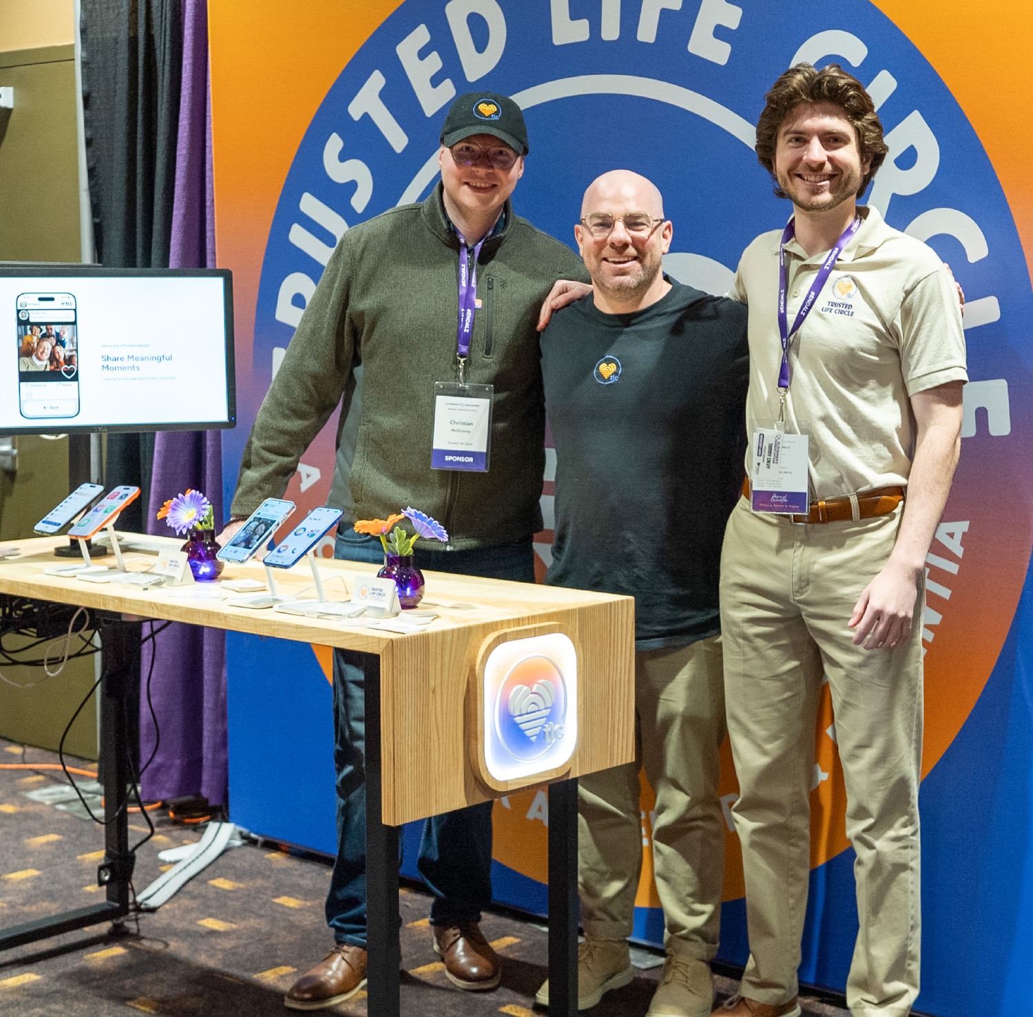 Three men pose at a conference booth with smartphones on a wooden table and a blue‑orange backdrop behind them.